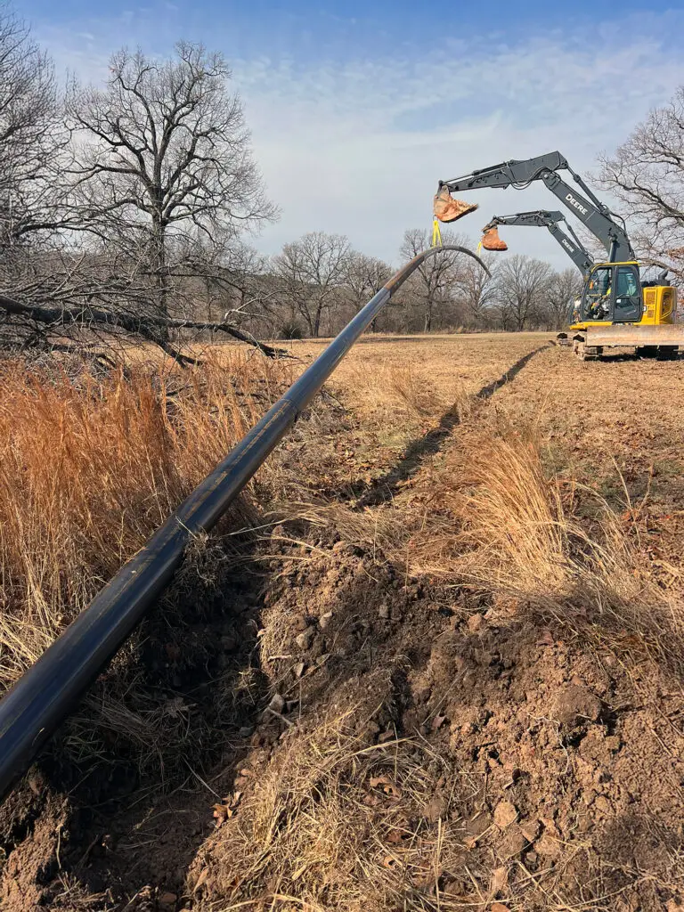 An excavator lifts and positions a long black conduit pipe across an open field, preparing it for burial. The pipe stretches from a freshly dug trench in the foreground toward the machinery, framed by dry grass, leafless trees, and a clear blue sky.